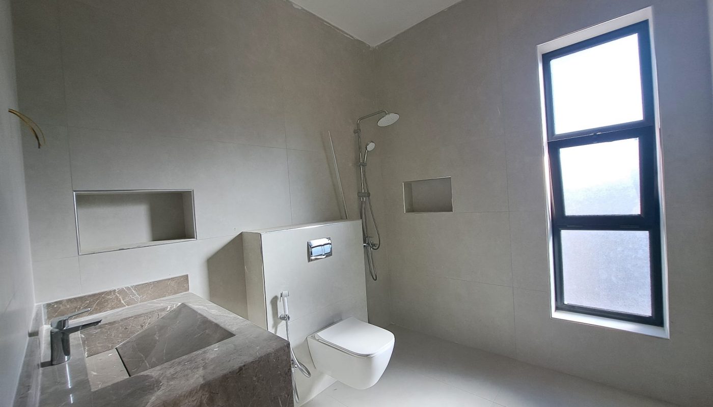 Modern bathroom with stone sink, wall-mounted toilet, walk-in shower, and a tall frosted window; neutral-toned tiles throughout.