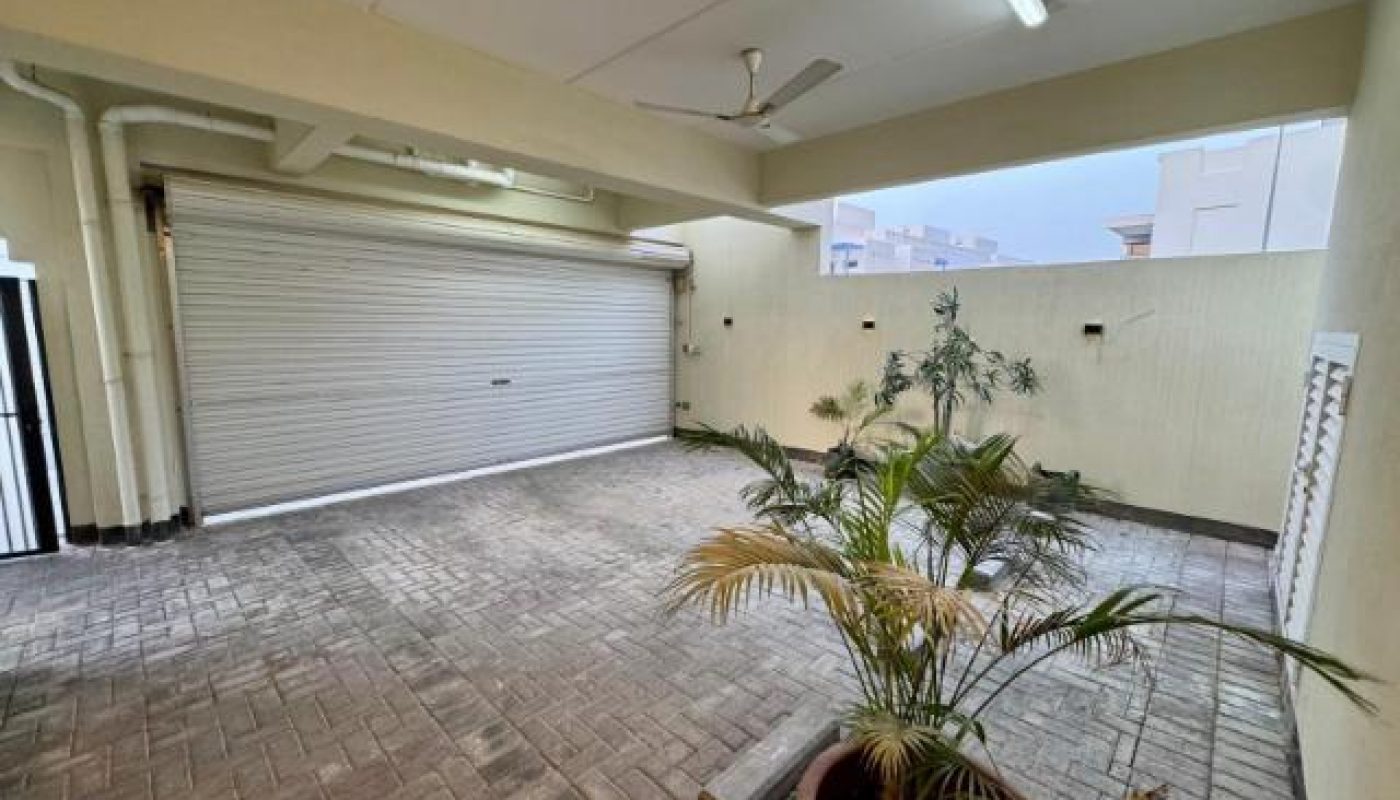 A covered garage with a closed metal rolling door, ceiling fan, fluorescent light, brick flooring, and several potted plants near the walls.