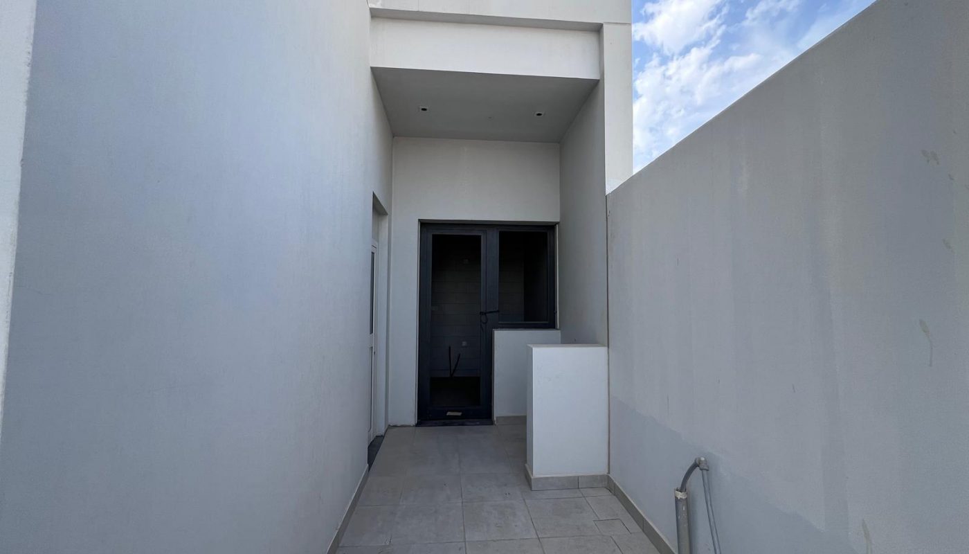 Narrow, tiled outdoor corridor with white walls, a recessed entry area at the end, and exposed pipes along the right side; blue sky with clouds above.