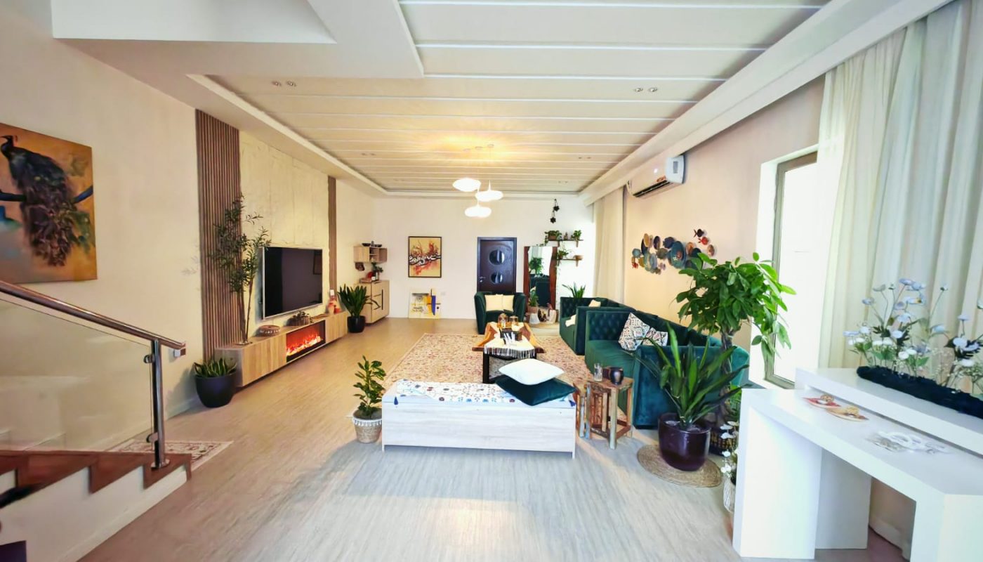 Modern living room with green sofa, indoor plants, wall art, TV, and a staircase on the left. Neutral tones and natural light create a spacious, inviting atmosphere.