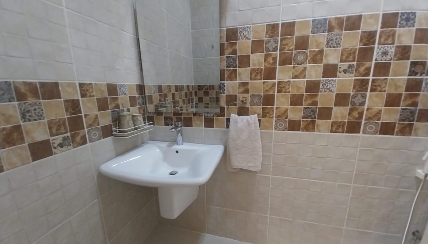 A bathroom sink with a mirror above, a towel hanging on a hook, and beige and brown patterned tiles on the wall.