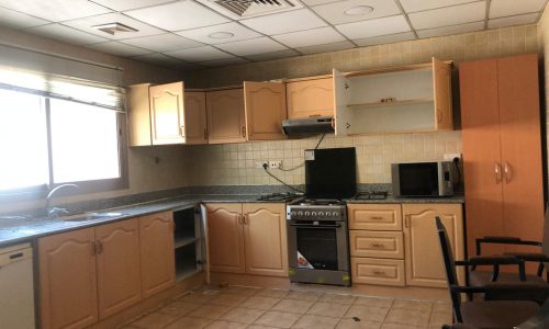 A kitchen with light wooden cabinets, some open, and granite countertops. It has a window, stove, microwave, dishwasher, and two chairs. Tile flooring and a drop ceiling with a vent are visible.