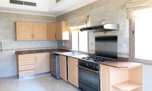 A modern kitchen with light wood cabinets, a stainless steel stove, dishwasher, sink, and granite countertops. The room features tiled walls and floor, with windows providing natural light.