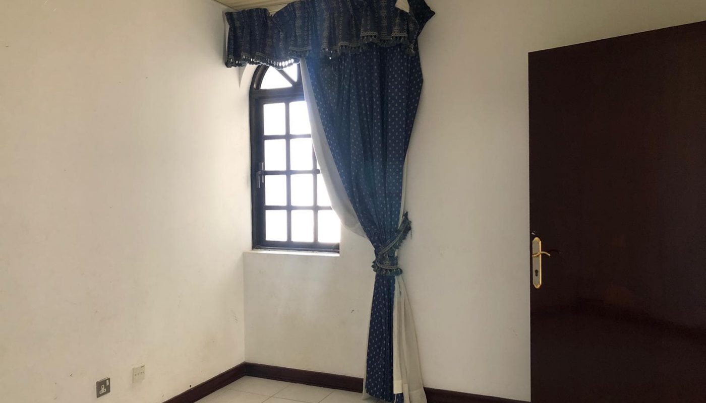 An empty room with a small window covered by blue curtains tied back, a white tiled floor, white walls, and a brown wooden door partially open.