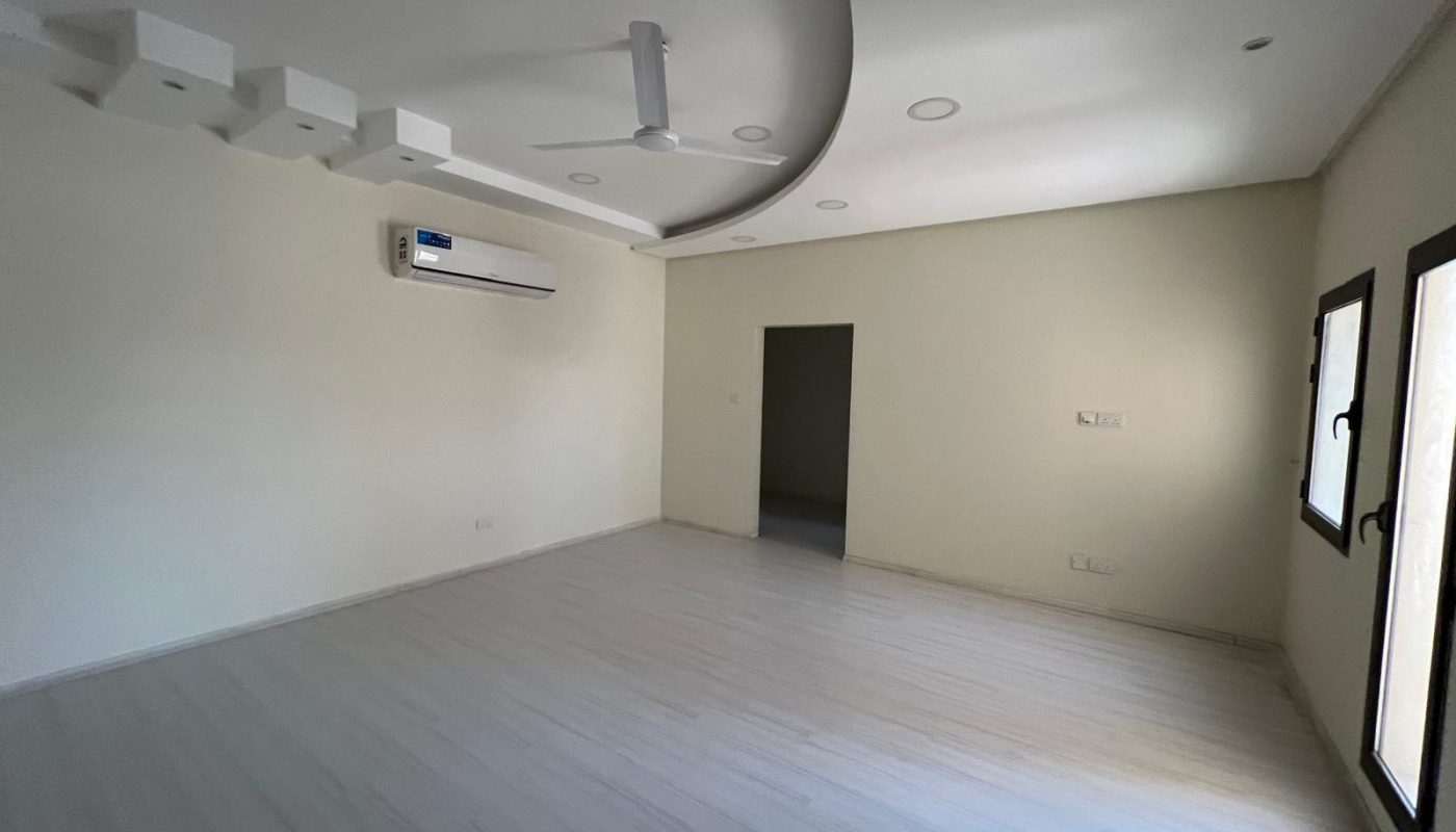 A beautiful villa for rent in Sanad features a vacant room with light-colored flooring, white walls, a ceiling fan, a wall-mounted air conditioner, and recessed lighting. Two windows on the right wall and a door leading to another room complete this serene space.