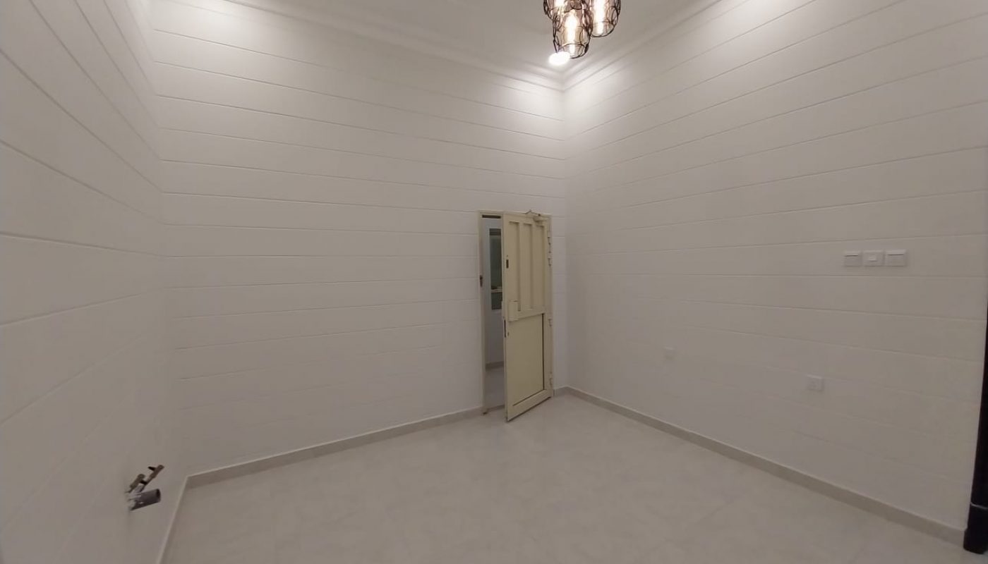Empty white room with tiled floor, panelled walls, ceiling lights, and a hanging light fixture. A door is open, revealing a hallway outside.