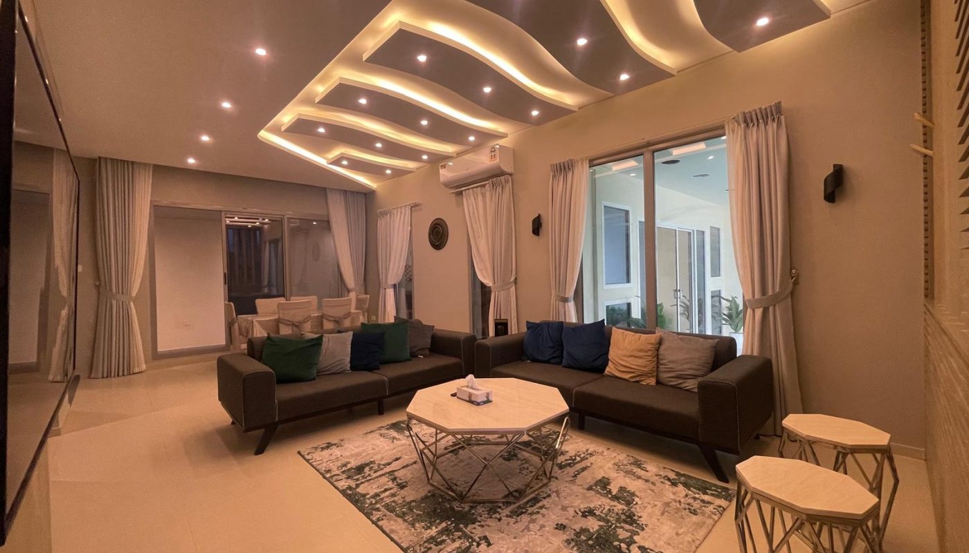 Modern living room with two dark sofas, a round coffee table, patterned rug, and decorative ceiling lights; large windows with curtains and a view to an outdoor area.