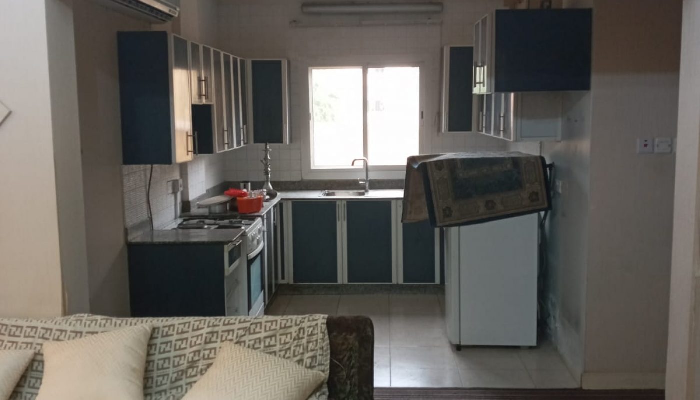 A small open kitchen with blue cabinets, a fridge, and a rug draped over it; a patterned sofa with cushions and children's toys is in the foreground.