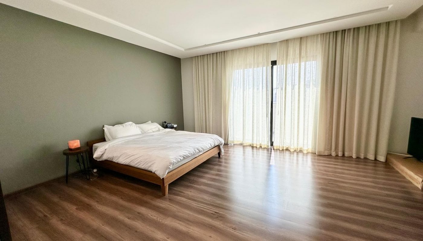 Minimalist bedroom with a low wooden bed, white bedding, small side table, and large window with sheer curtains. Room features wood flooring and neutral-colored walls.
