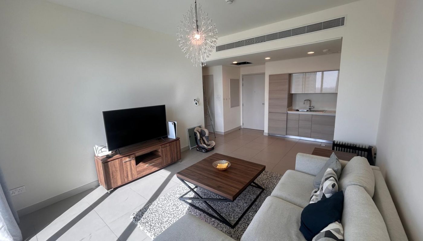 Modern living room with a gray sectional sofa, wooden coffee table, TV on a stand, and open kitchen in the background. Room is well-lit by natural light.