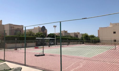 A fenced outdoor tennis court in a residential area with several low-rise buildings and greenery in the background under a clear blue sky.