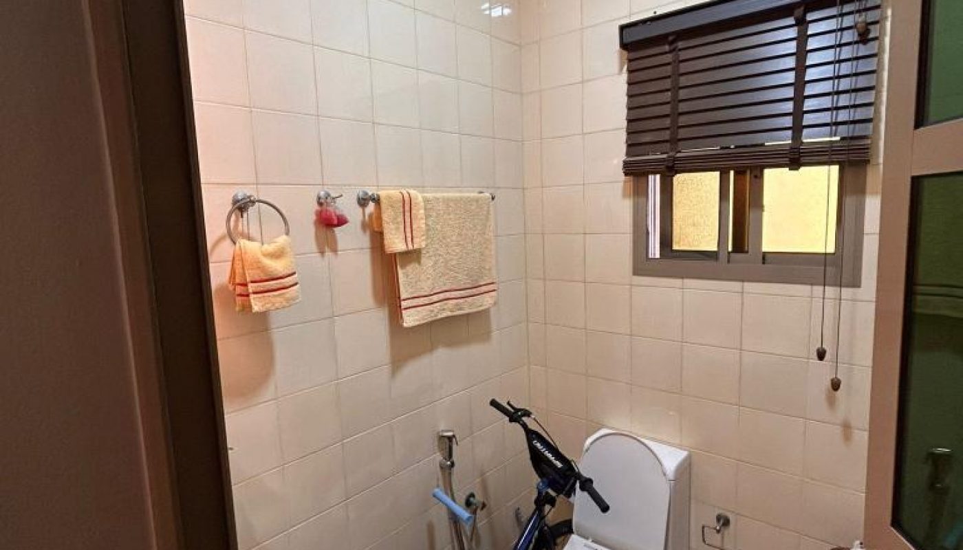 Small bathroom with cream tiles, a sink, a toilet, a towel rack with towels, a window with blinds, and an exercise bike placed between the toilet and the sink.