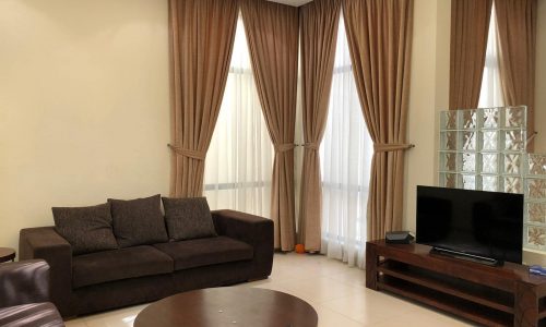 A living room with brown curtains, a dark brown sofa, a round wooden coffee table, and a wooden TV stand with a flat-screen TV. Bright neutral walls and large windows add light to the space.