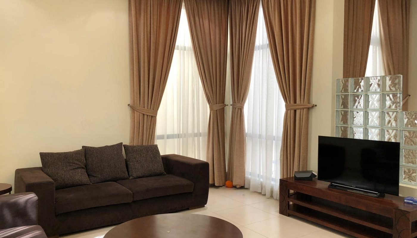 A living room with brown curtains, a dark brown sofa, a round wooden coffee table, and a wooden TV stand with a flat-screen TV. Bright neutral walls and large windows add light to the space.