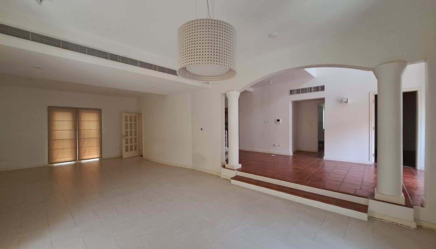Spacious empty room with white walls, tiled floors, a large ceiling light, arched entryway, and tall windows with closed blinds.