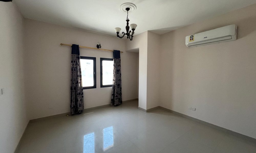 A bright, empty room with tiled flooring, a window with patterned curtains, a ceiling light fixture, and an air conditioner mounted on the wall.