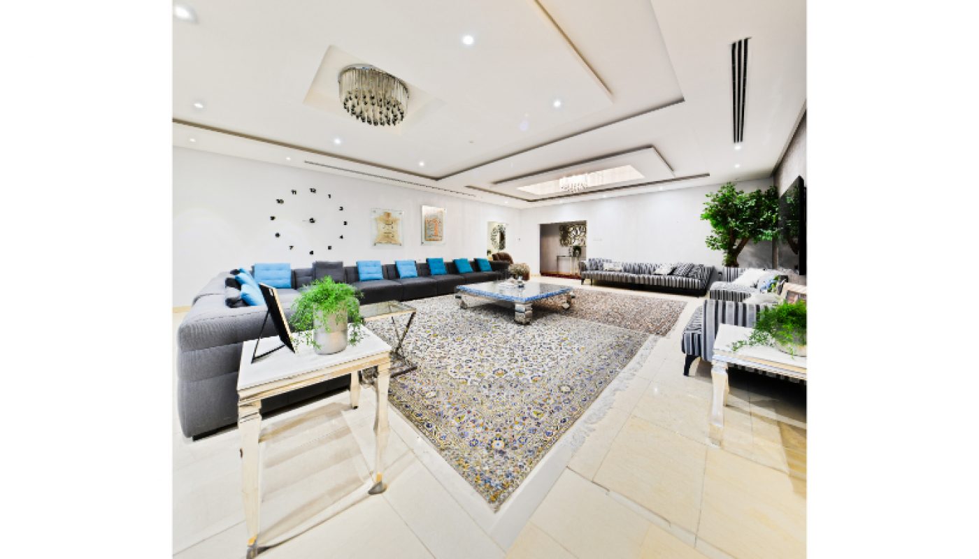 Spacious living room with two large gray sectional sofas, blue accent pillows, a patterned area rug, coffee tables, wall art, and indoor plants under recessed lighting.