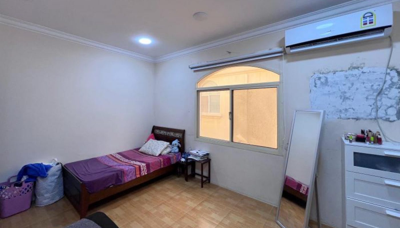 A simple bedroom with a single bed, a window with blinds, a wall-mounted air conditioner, a dresser with a mirror, and various personal items on the floor and furniture.