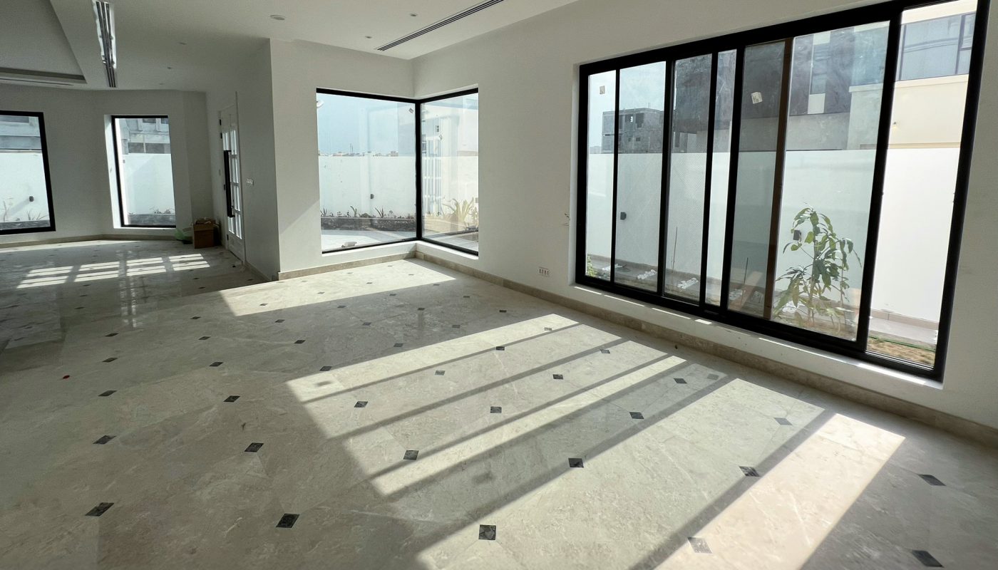 Spacious, empty room with large windows, tiled floor, and sunlight streaming in; white walls and view of an outdoor area through the glass.