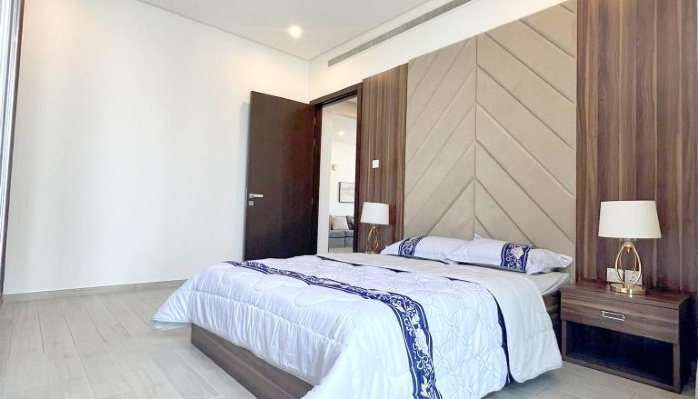 A modern bedroom with a double bed, two bedside tables with lamps, light wood flooring, and an open door leading to another room.