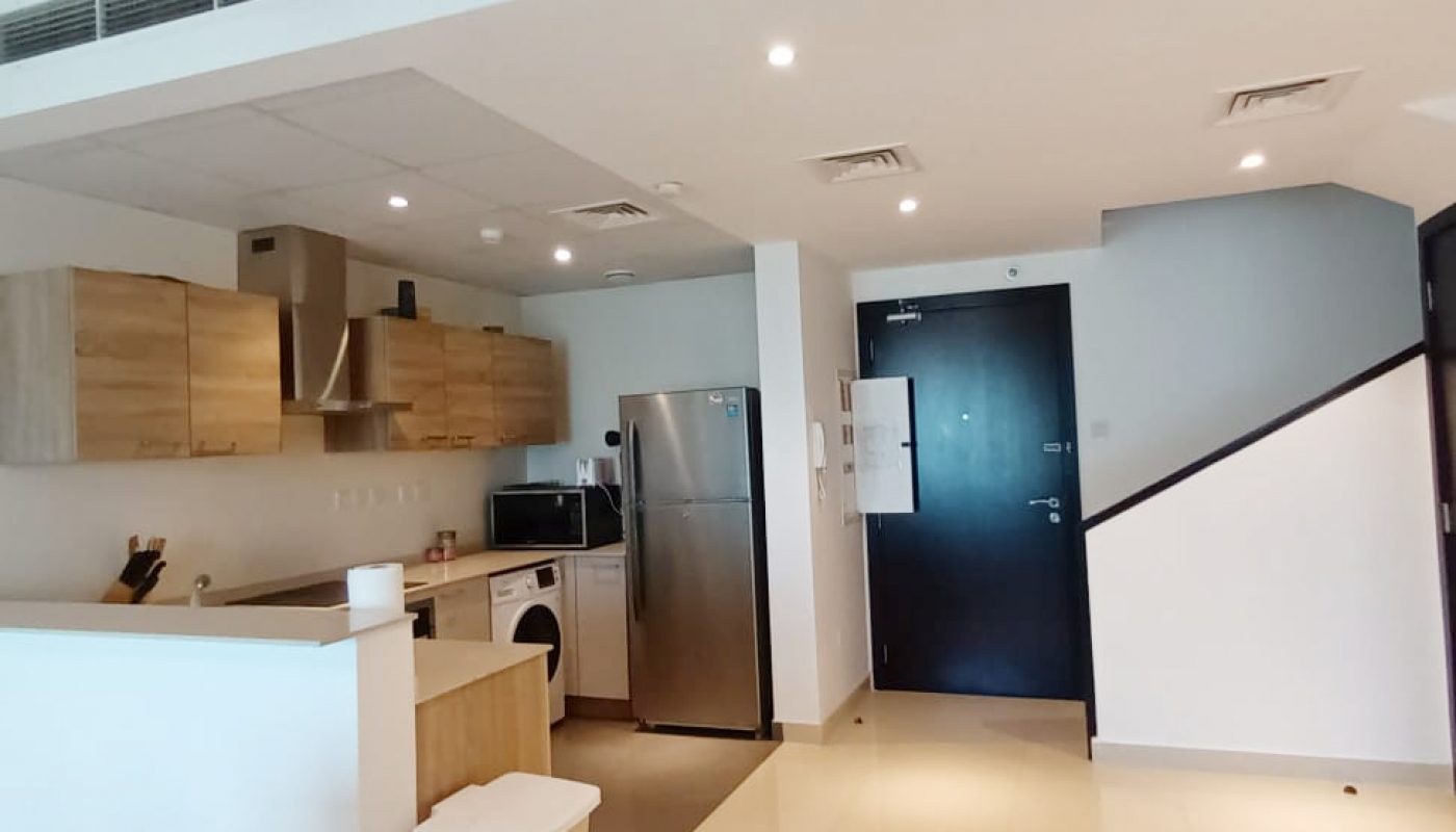 Modern kitchen with light wood cabinets, stainless steel appliances, and a white countertop next to an open entryway with a black door and tiled floor.
