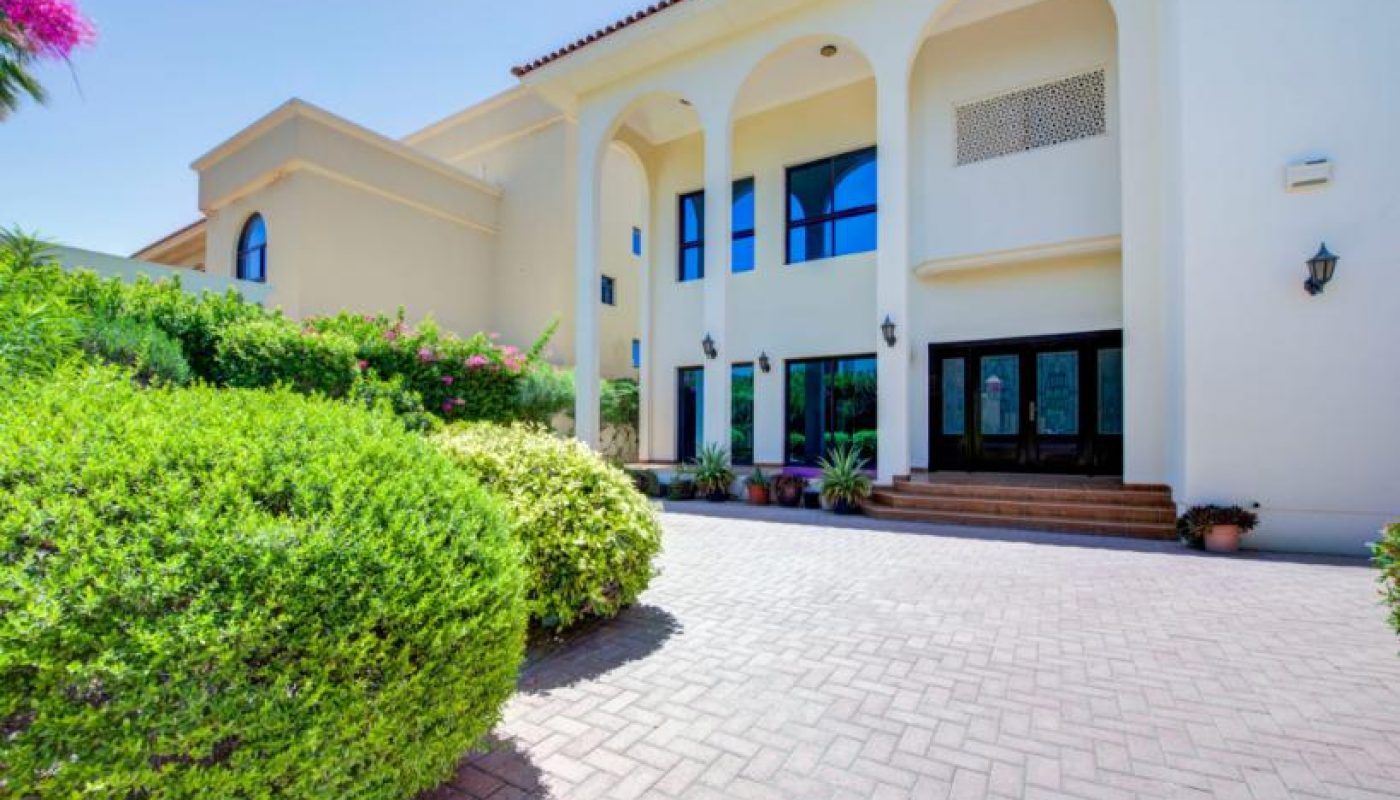 Large modern house with cream exterior, arched pillars, and multiple windows, surrounded by manicured bushes under a clear blue sky.