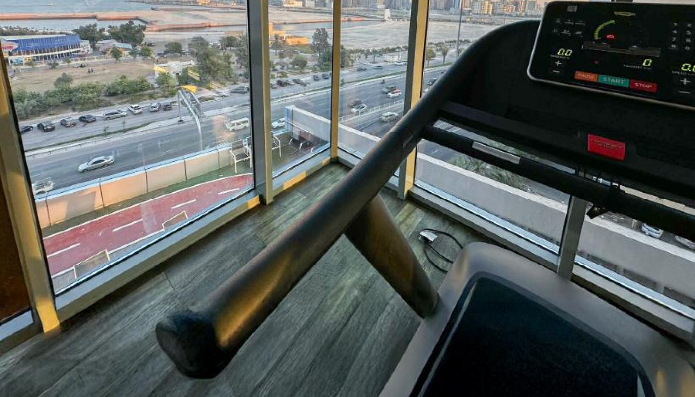 Treadmill facing large windows with a cityscape and waterfront view at sunset, overlooking roads and tall buildings.