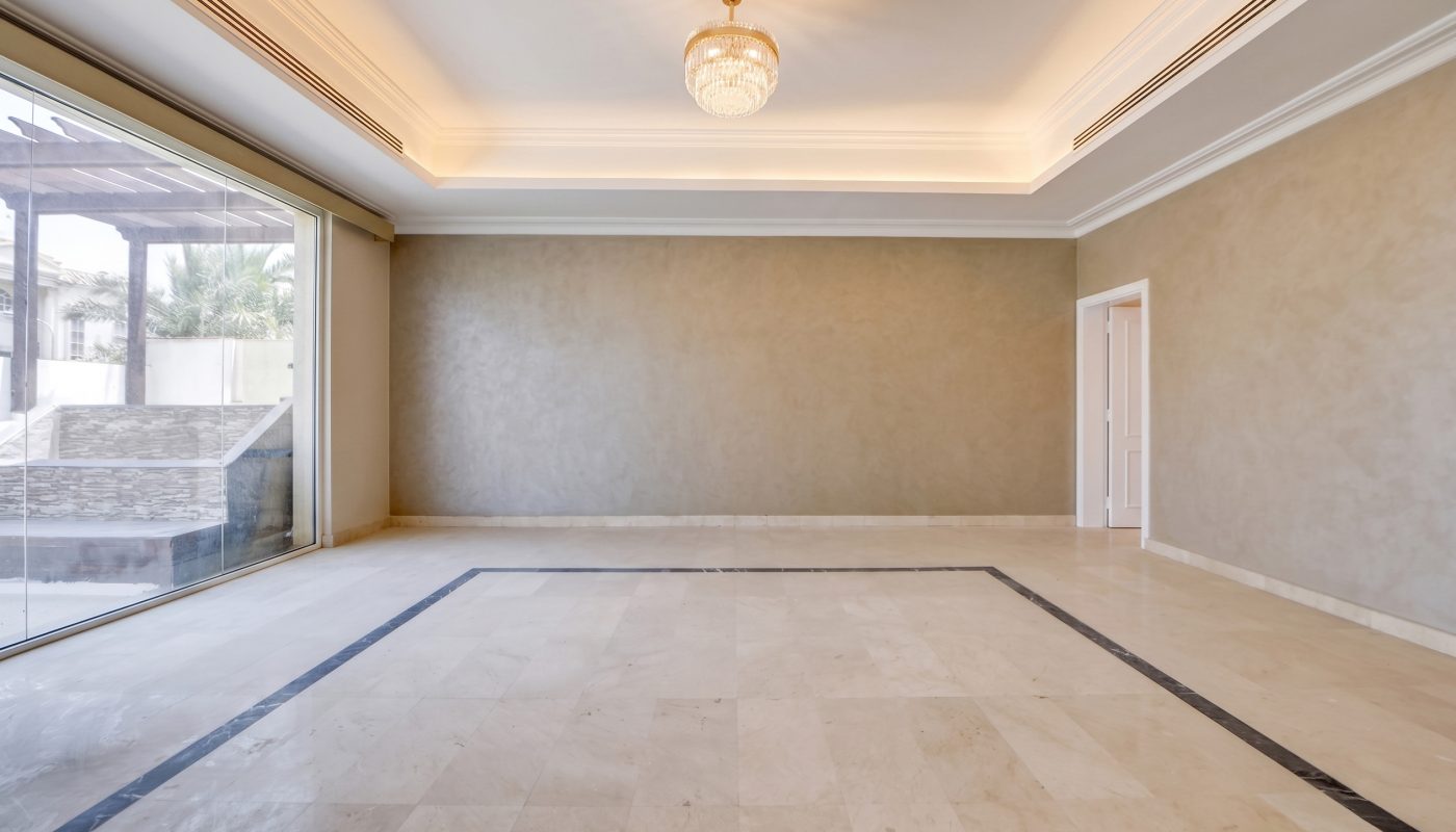 Empty room with beige marble flooring, plain taupe walls, a large window, recessed ceiling lights, and a decorative ceiling light fixture. Doorway and outdoor view visible on the left.