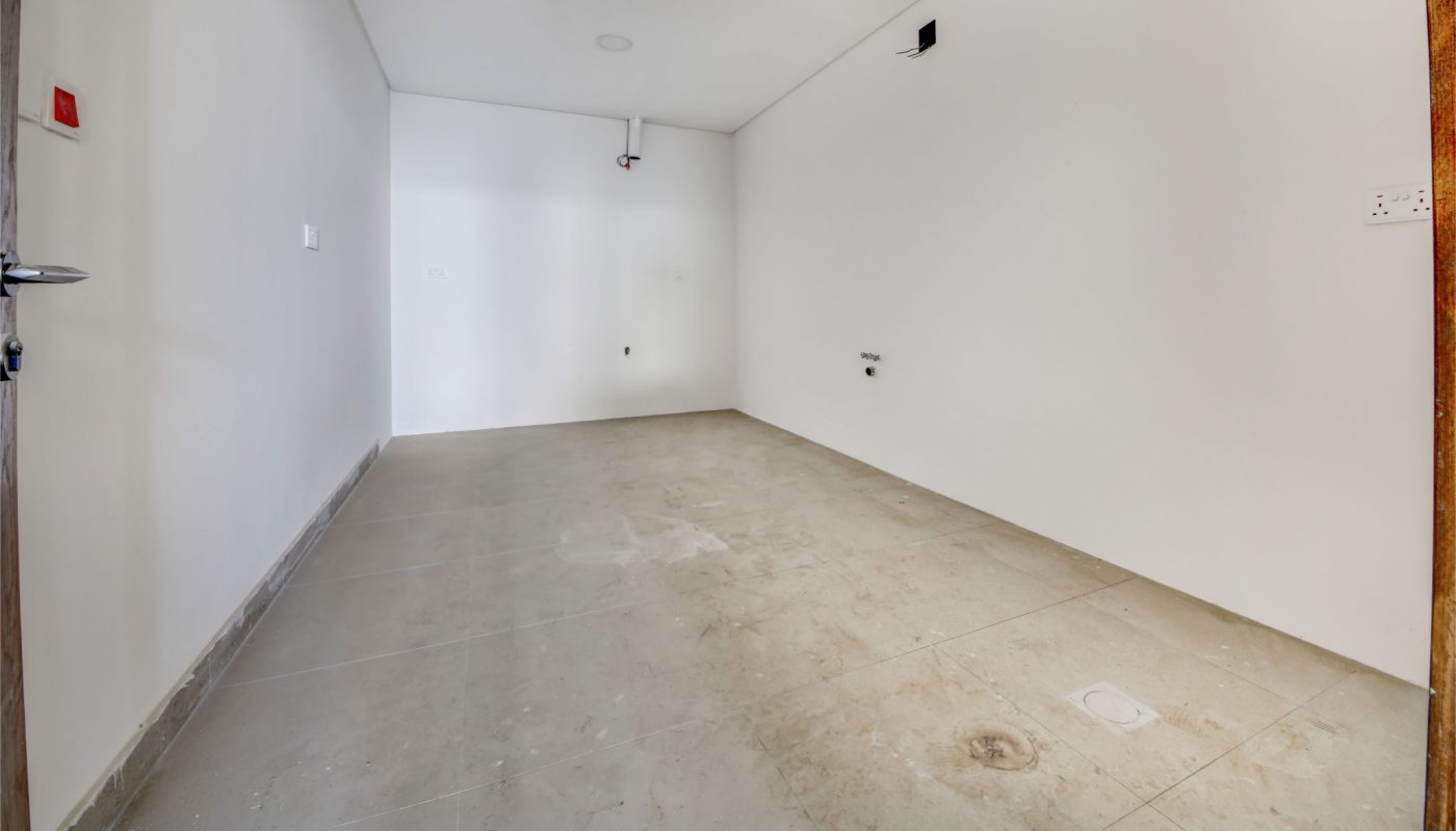 Empty, unfurnished room with white walls, a brown door, and a bare concrete floor. Ceiling-mounted fixtures and wall outlets are visible. No windows or furniture.