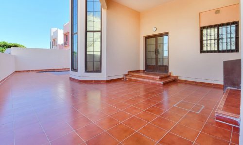 Spacious outdoor area with red-tiled flooring and beige walls, featuring a staircase leading to glass-paneled doors and large windows in this elegant 3BR villa in Al Areen.