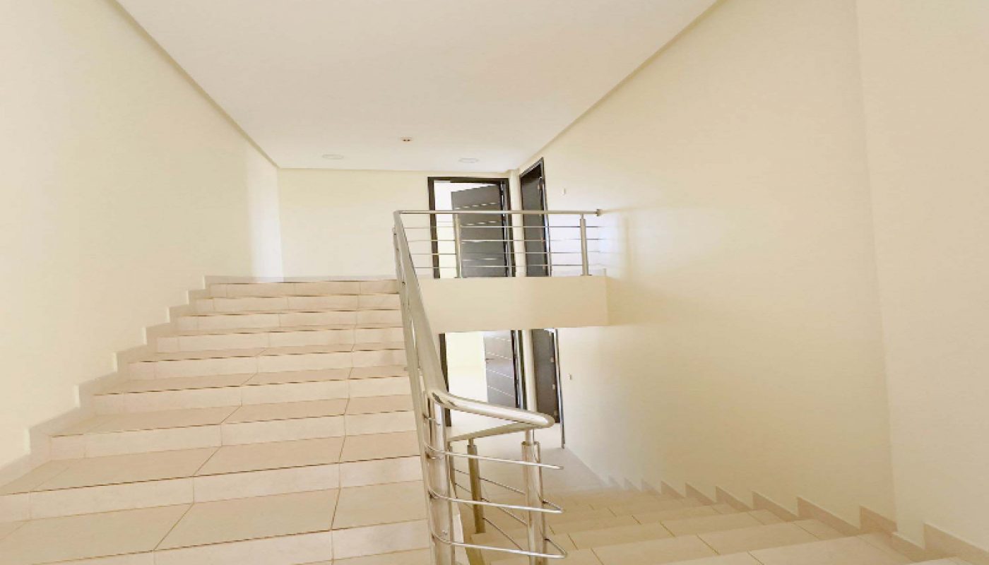 Indoor staircase with beige tiles and metal handrails, leading both upstairs and downstairs in a well-lit, neutral hallway.