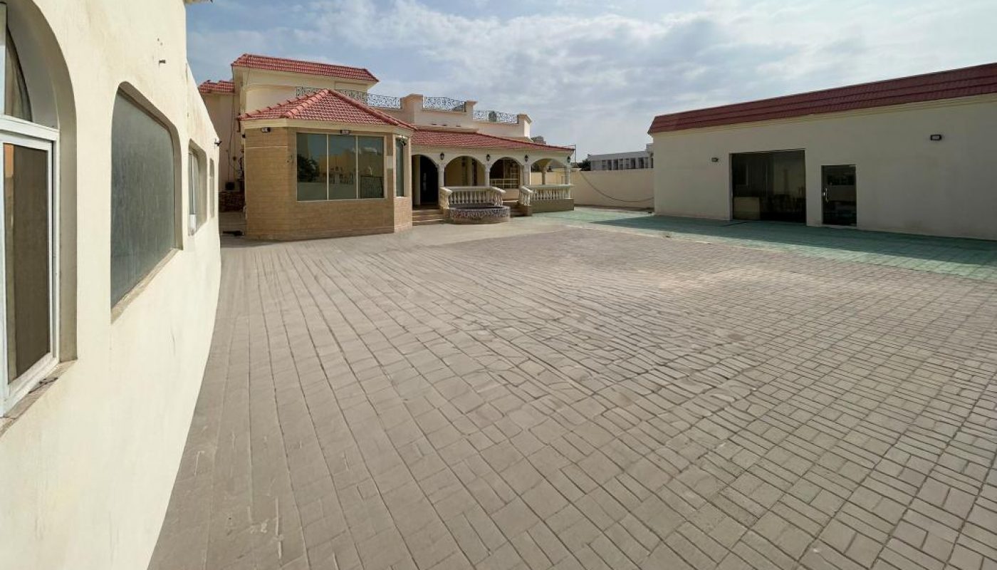 A wide, paved courtyard with a building on the left, a single-story structure with arches ahead, and another building with an open garage door on the right under a bright sky.