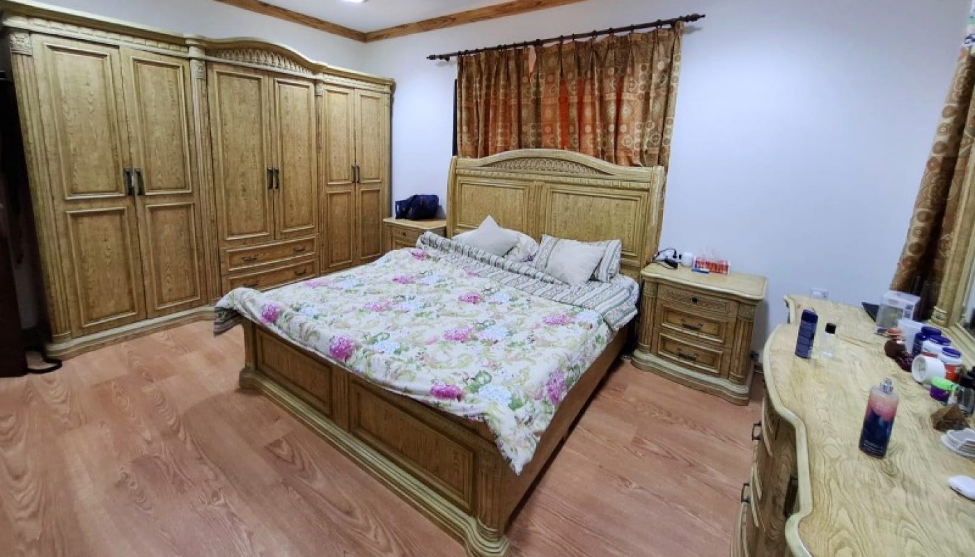 Spacious bedroom with wooden furniture, a double bed with floral bedding, large wardrobe, dresser, nightstands, and curtains over the windows. Ceiling fan and bright lighting fixtures overhead.