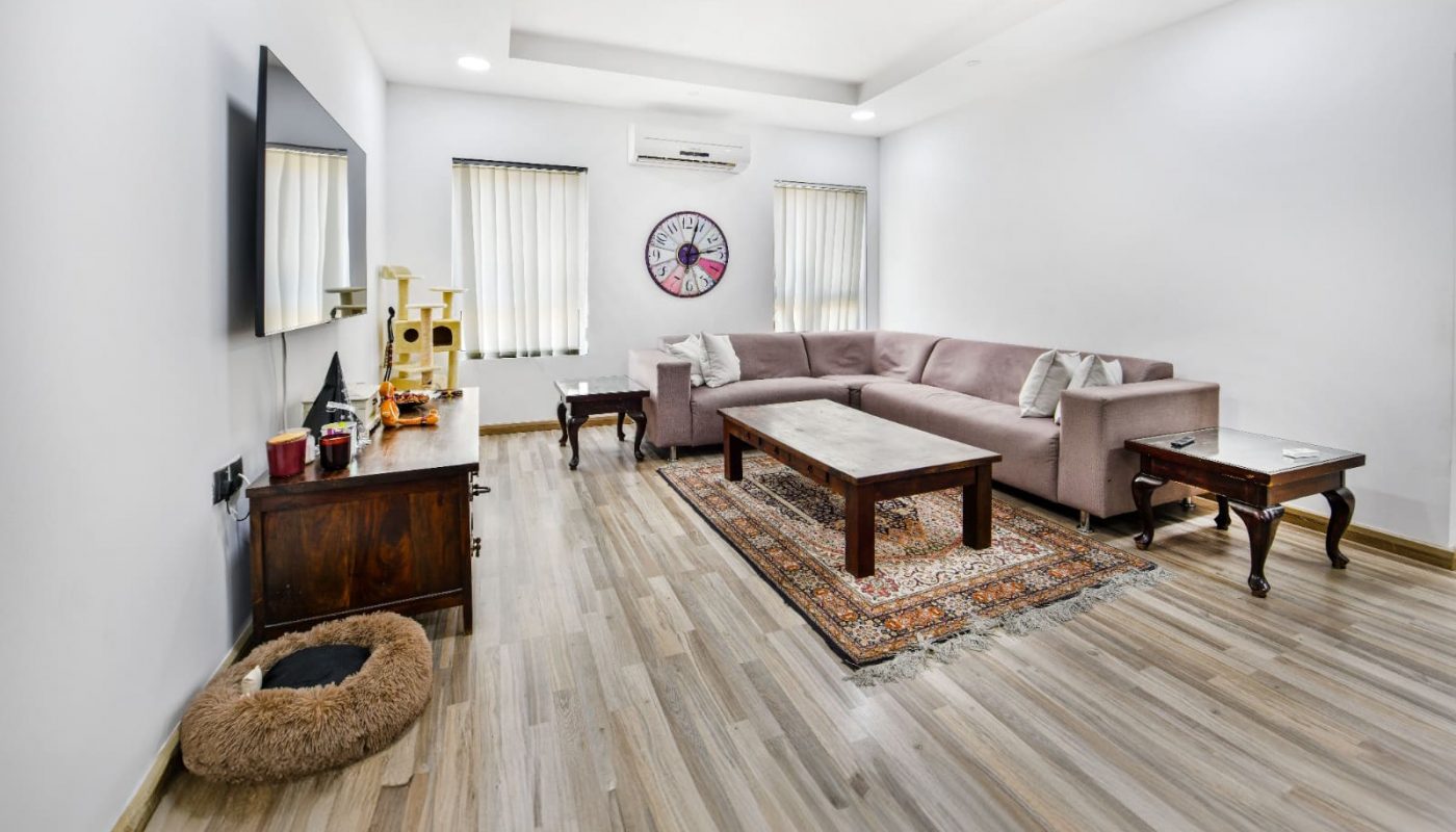 Spacious living room with a U-shaped sofa, wooden coffee table on a rug, wall-mounted TV, and a cat bed on the floor. Two narrow windows are seen, and a clock decorates the wall.