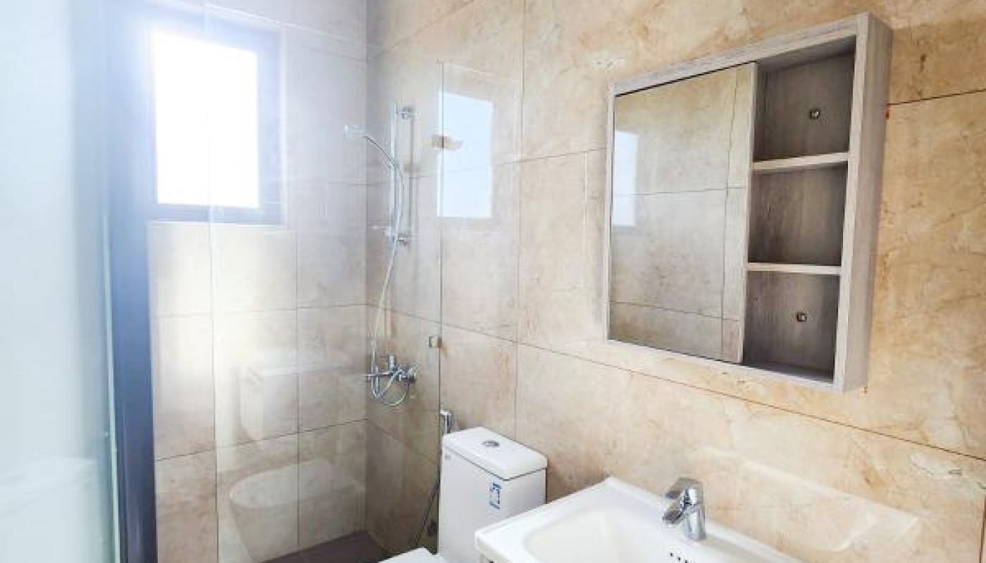 Modern bathroom with beige tiled walls, toilet, white sink vanity, wall-mounted mirror cabinet, and a shower area by a frosted window.