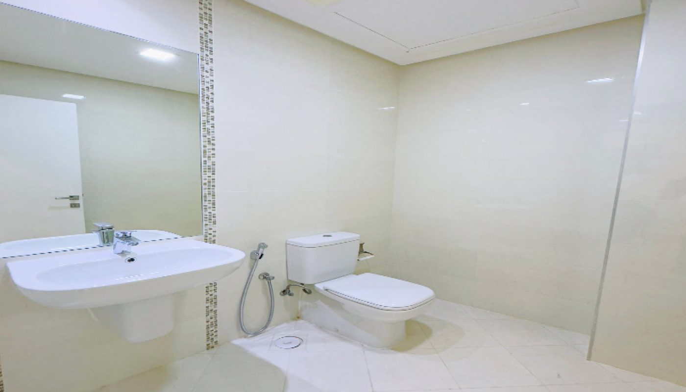 A clean, modern bathroom with a wall-mounted sink, a toilet, a handheld bidet sprayer, and cream-colored tiled walls and floor.