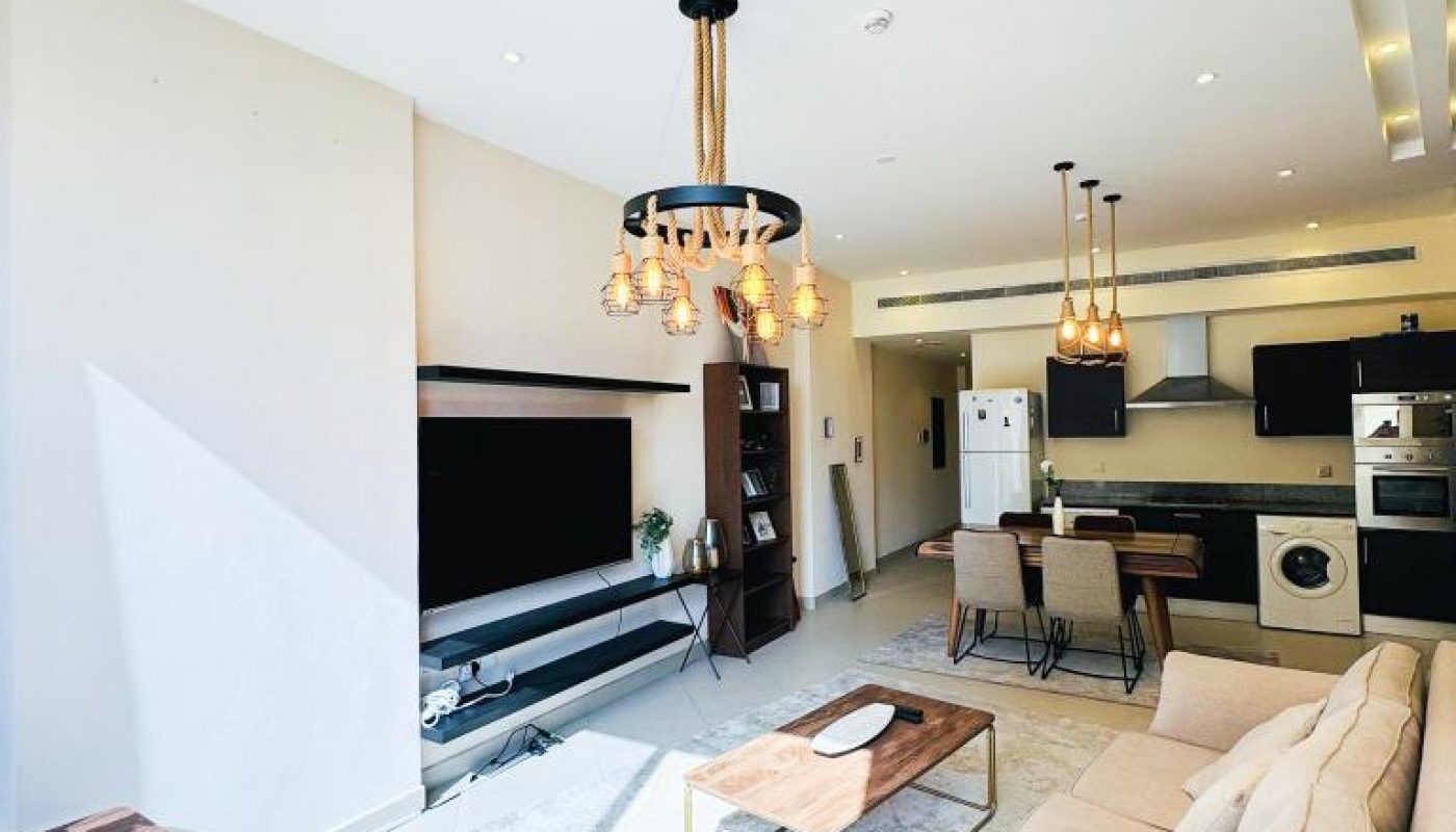 Modern open-plan living room and kitchen with beige sofa, wall-mounted TV, dining table, and contemporary lighting fixtures. Natural light fills the space.