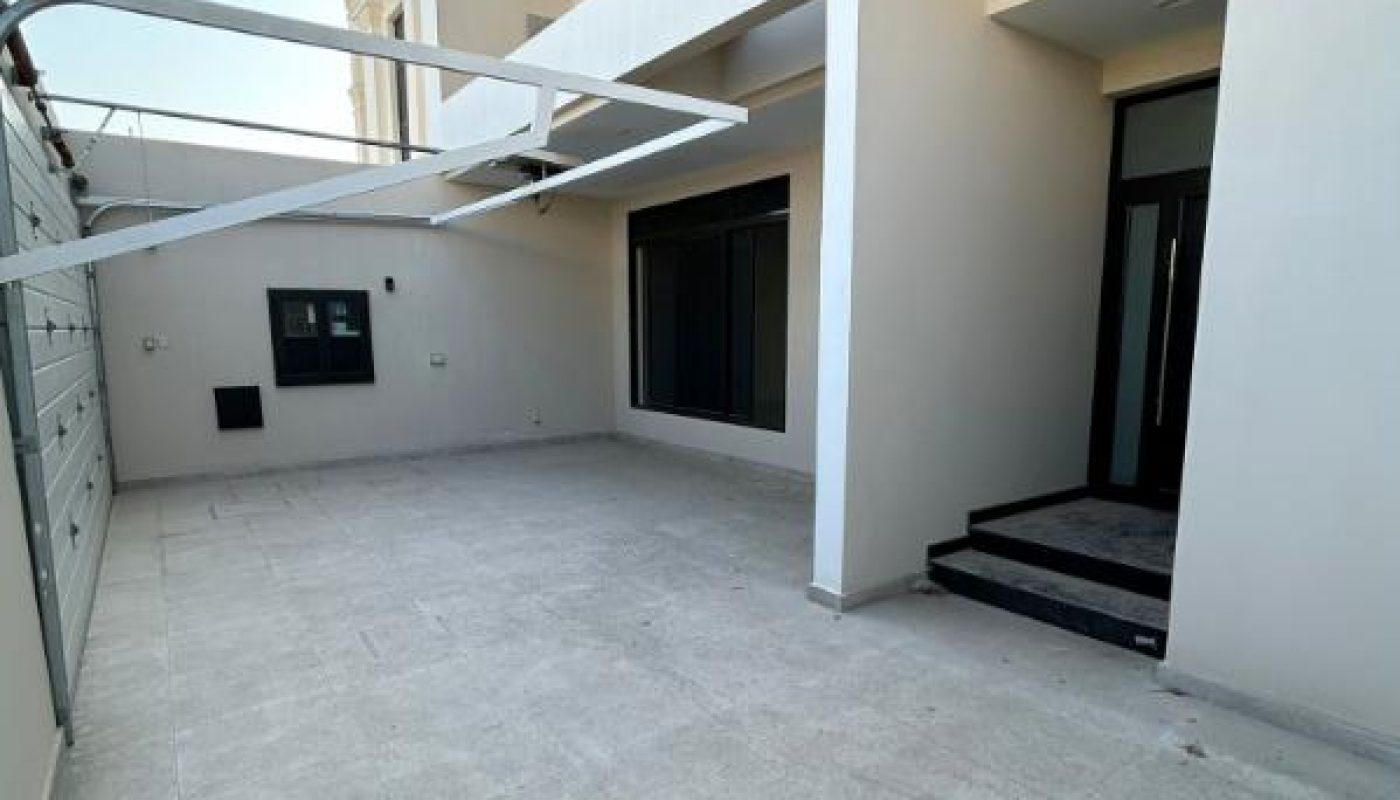 Modern, two-story house exterior with a spacious tiled patio, large windows, steps leading to a glass door, and a partial pergola frame overhead.