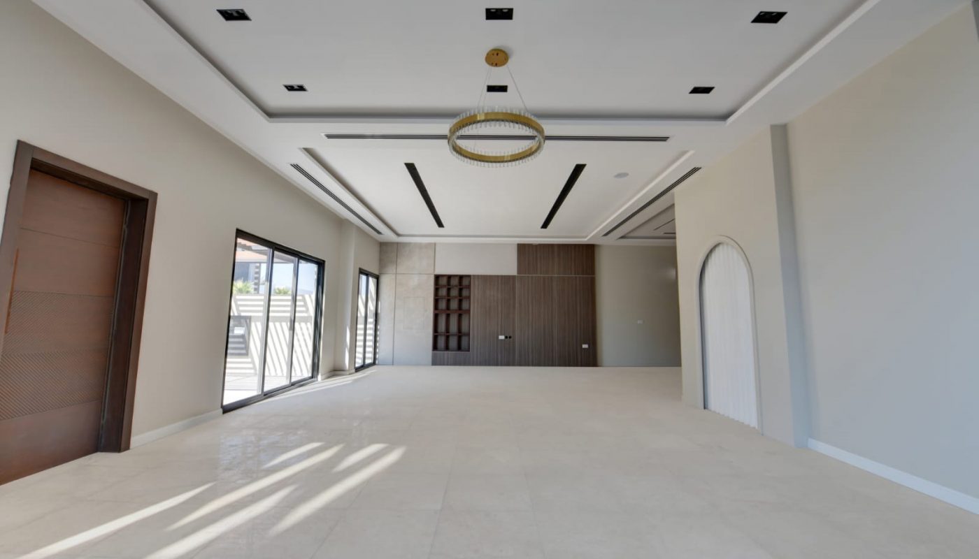 Spacious, empty modern room with large tile floor, recessed lighting, chandelier, wood-paneled accent wall, and sliding glass doors letting in natural light.