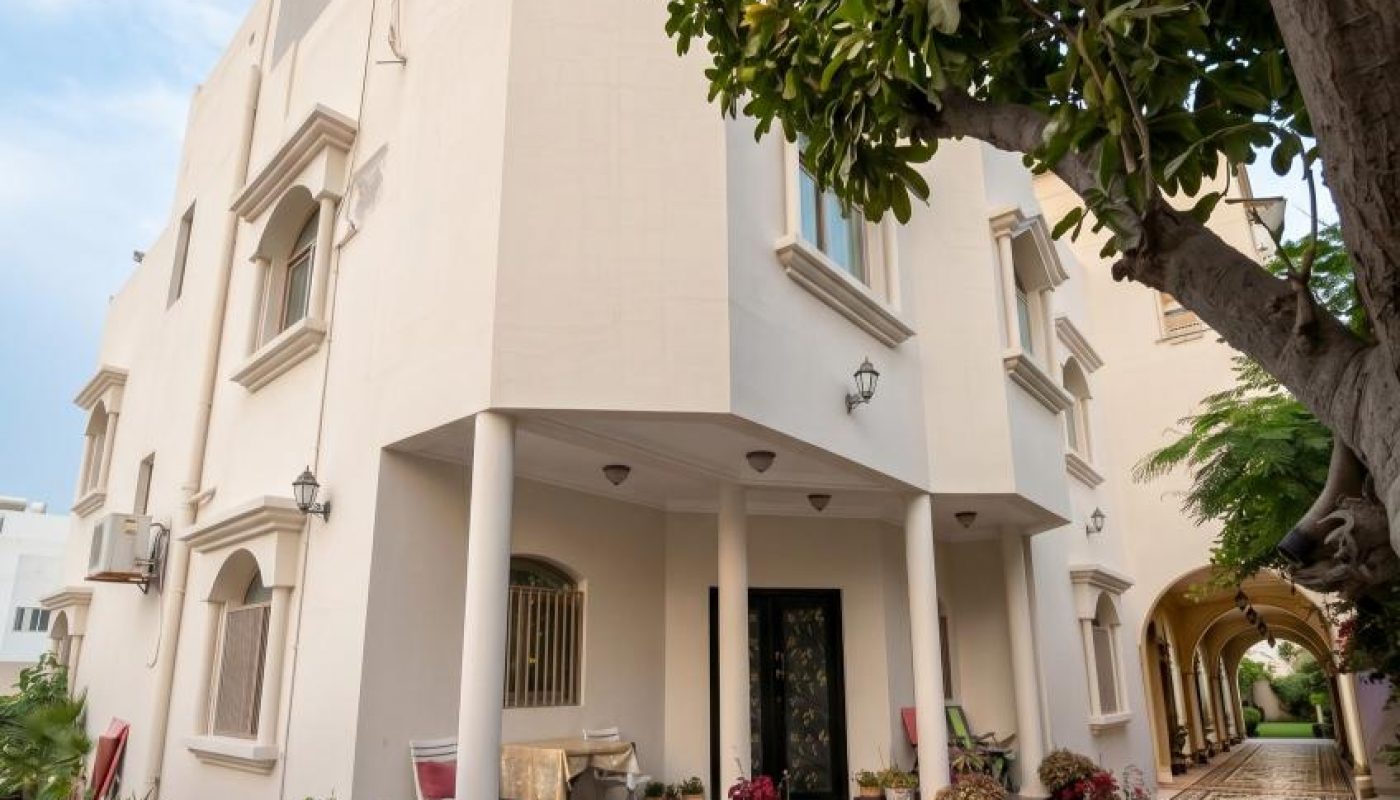 A two-story cream-colored house with arched windows and a front porch, surrounded by potted plants and trees on a tiled pathway.