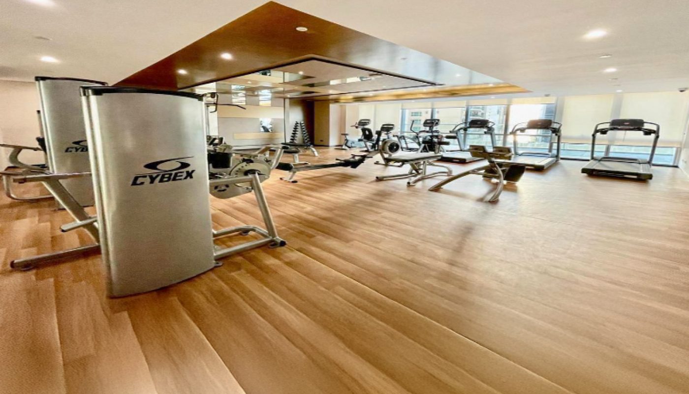 Spacious gym with wooden floors, exercise machines including treadmills, stationary bikes, and weight equipment, and large windows letting in natural light.