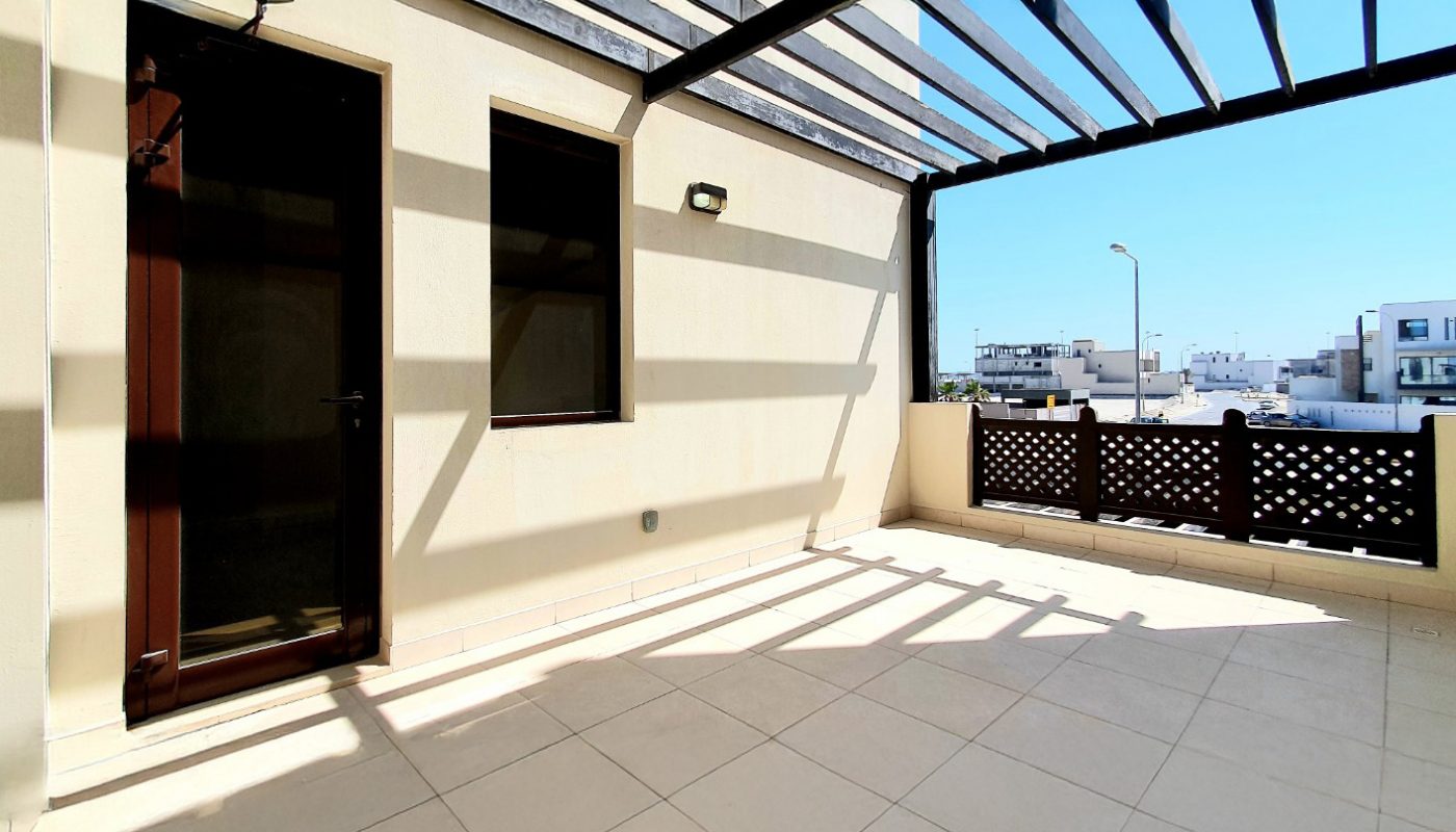 Sunny outdoor terrace with tiled floor, partial roof pergola casting shadows, a brown door and window on the left, and a patterned railing overlooking other buildings.