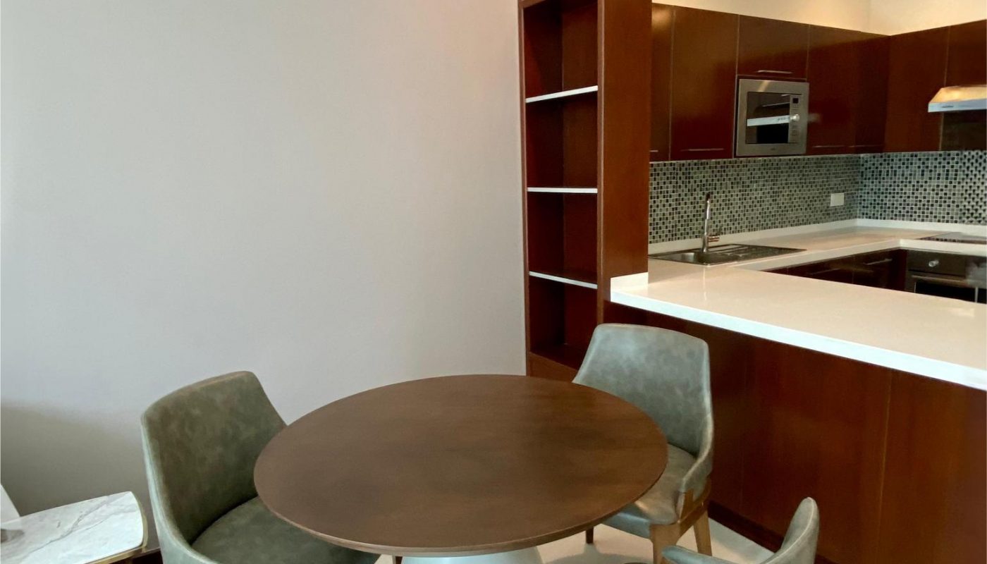 A small round dining table with three gray upholstered chairs sits near a modern kitchen with wood cabinets and white countertops.