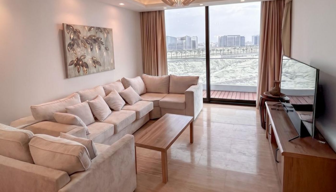 Modern living room with a sectional sofa, wooden coffee table, wall art, TV on a console, and large windows offering a city and water view.
