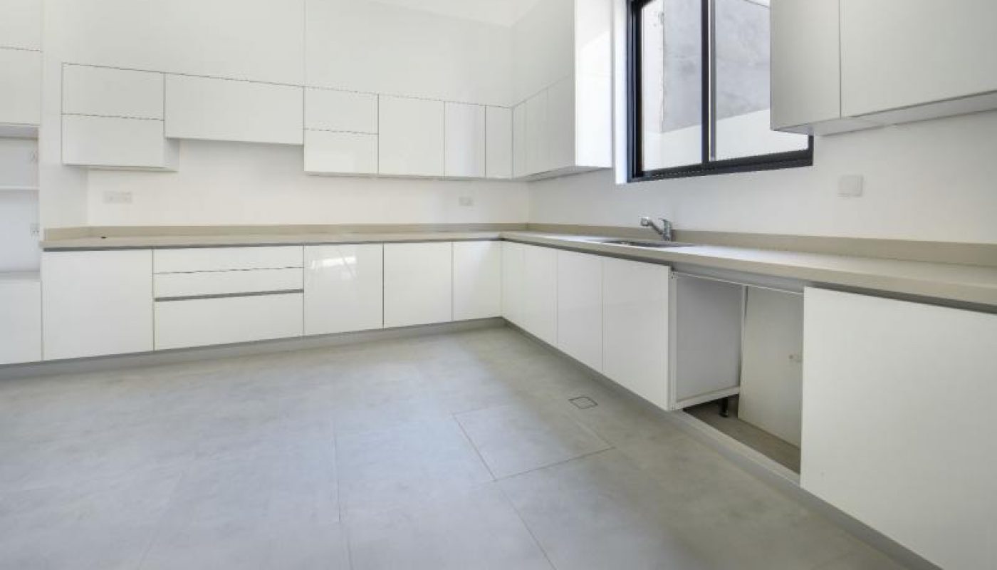 Modern kitchen with white cabinets, light countertops, large floor tiles, and a window above the sink. One cabinet door is open, revealing empty space underneath.