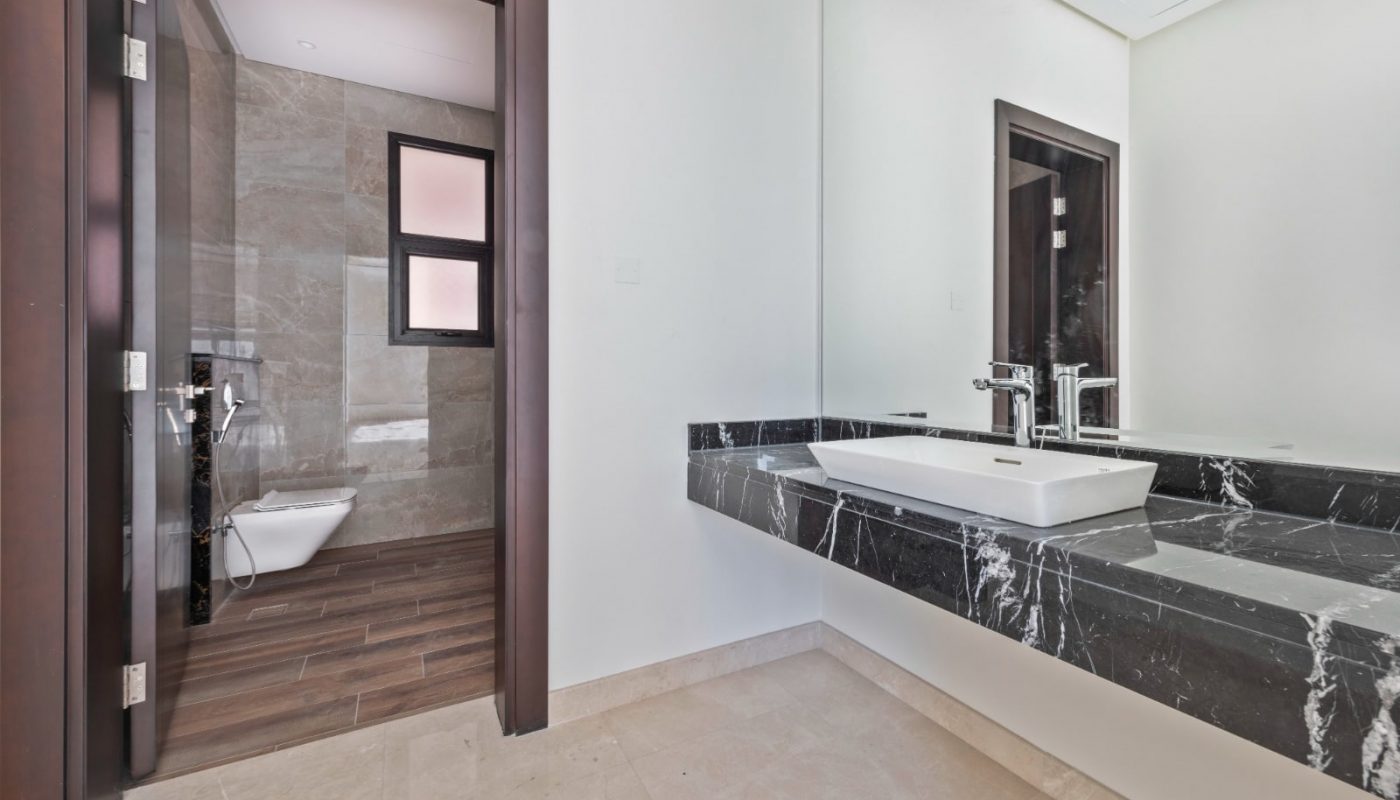 Modern bathroom with beige tile floor, large mirror above a black marble sink, and visible entrance to a separate toilet space with a window on the left.