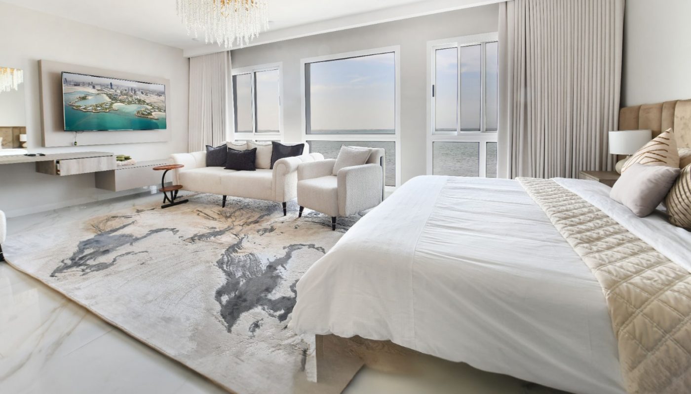 Spacious, modern bedroom with a large bed, white and beige decor, abstract rug, a wall-mounted TV, chandelier, and windows overlooking the ocean.