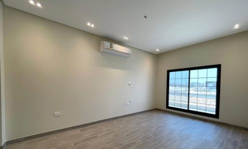 This elegant villa in Diyar Al Muharraq features a spacious, empty room with wooden floors, white walls, ceiling lights, and a large window with sliding glass doors. A wall-mounted air conditioning unit is positioned above the window. This beautiful property is now for sale.