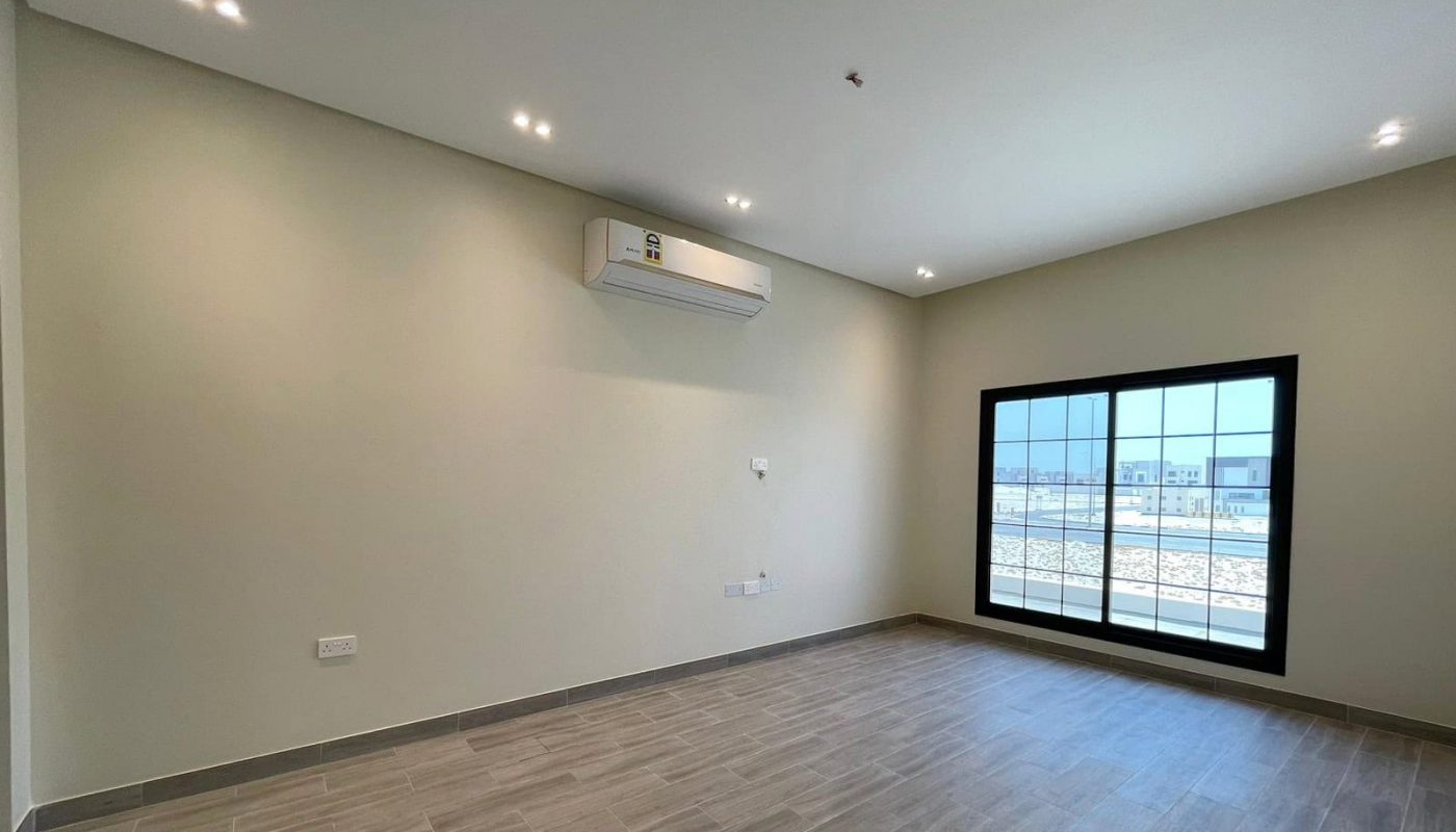 This elegant villa in Diyar Al Muharraq features a spacious, empty room with wooden floors, white walls, ceiling lights, and a large window with sliding glass doors. A wall-mounted air conditioning unit is positioned above the window. This beautiful property is now for sale.