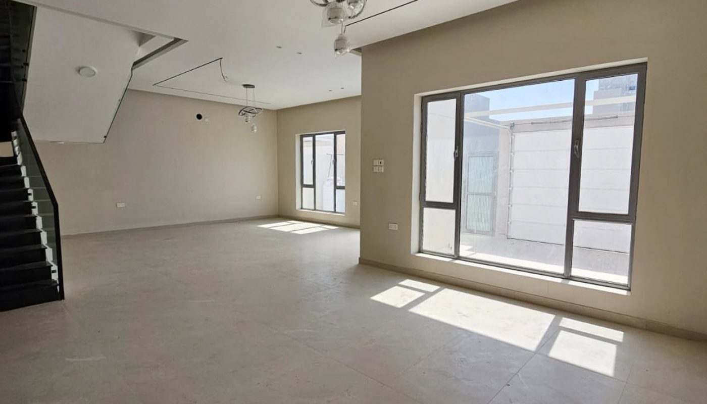 Spacious, unfurnished living room with beige walls, large windows, and tiled floor. A modern staircase and ceiling light fixtures are visible.